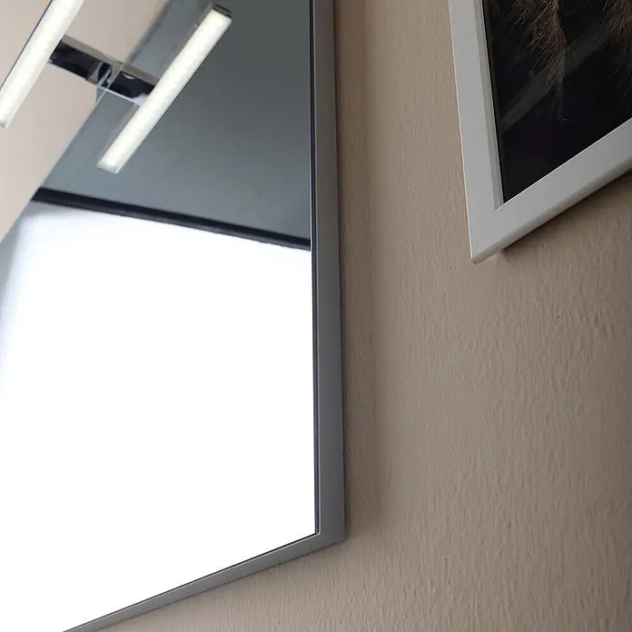 Kiamami Valentina Badmöbel-Set - Grau - Rechteckiger Spiegel mit silbernem Rahmen und LED-Beleuchtung, an einer strukturierten Wand neben einem weißen Bilderrahmen.