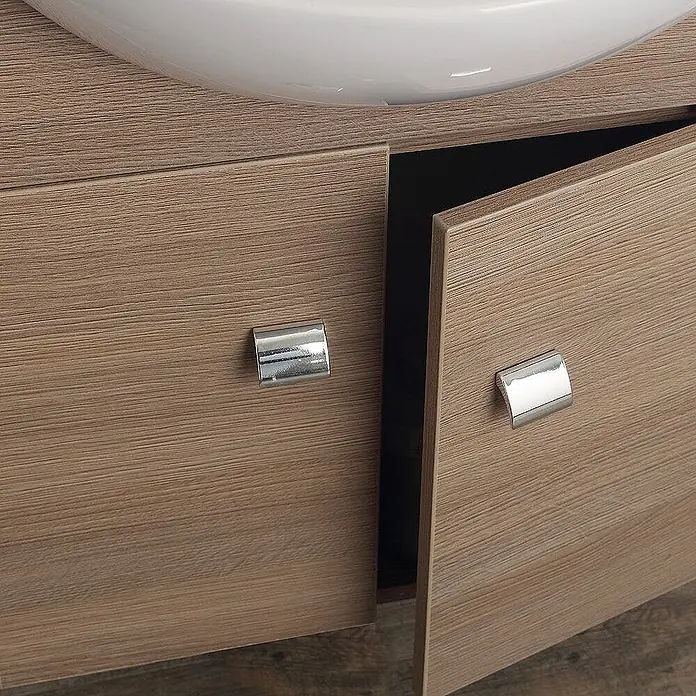 Kiamami Valentina Badmöbel-Set - Helles Holz - Heller Holzmaserung-Badezimmerschrank mit zwei leicht geöffneten Türen und Chromgriffen, unter einem weißen Waschbecken.