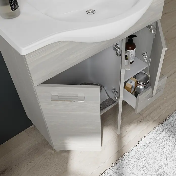 Kiamami Valentina Badmöbel-Set - Helles Holz - Helle Badezimmereinheit in Holzoptik mit weißem Waschbecken und geöffneten Türen, die Pflegeprodukte und ein Handtuch zeigen.