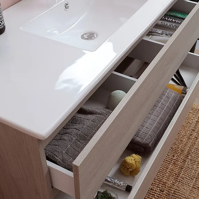 Kiamami Valentina Badmöbel-Set - Helles Holz - Waschtischunterschrank in heller Holzoptik mit integriertem weißen Waschbecken und offenen Schubladen, die Handtücher und Toilettenartikel zeigen.