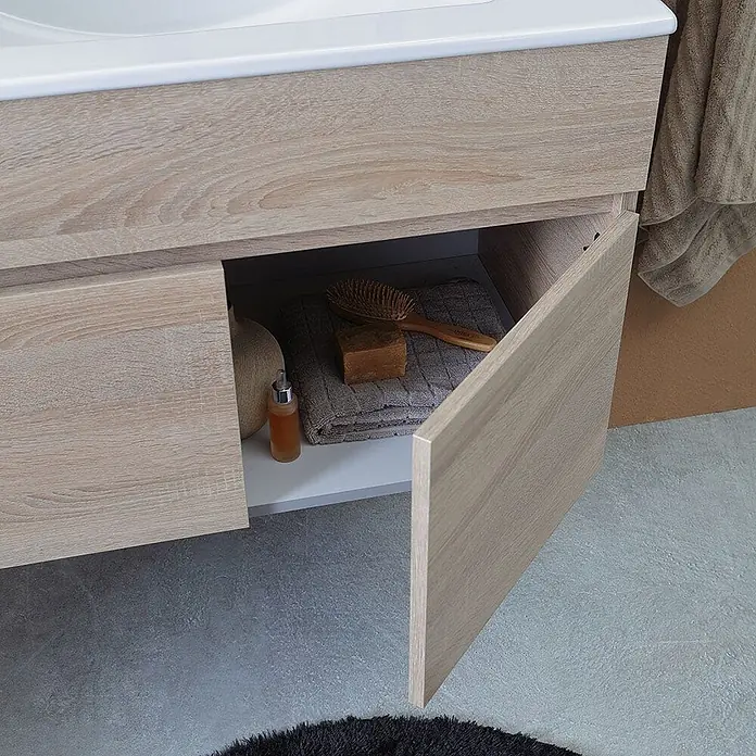 Kiamami Valentina Badmöbel-Set - Braun - Waschtischunterschrank aus heller Holzmaserung mit offener Tür, die Handtuch, Bürste, Seife und Flasche zeigt.