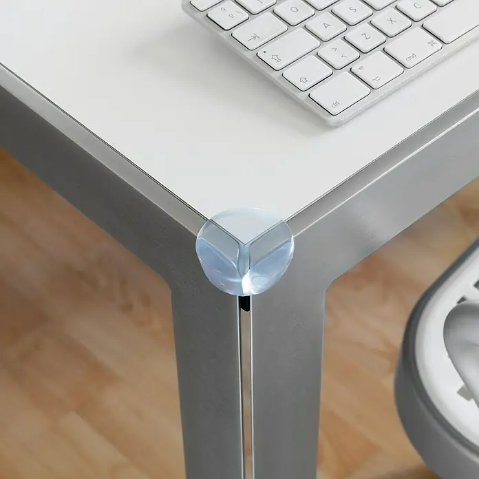 Inofix Protector para esquina 5000-0- - Transparente, 4 ud. - Primer plano de un protector de esquina transparente en un borde de mesa para proteger contra golpes, con parte de un teclado visible.