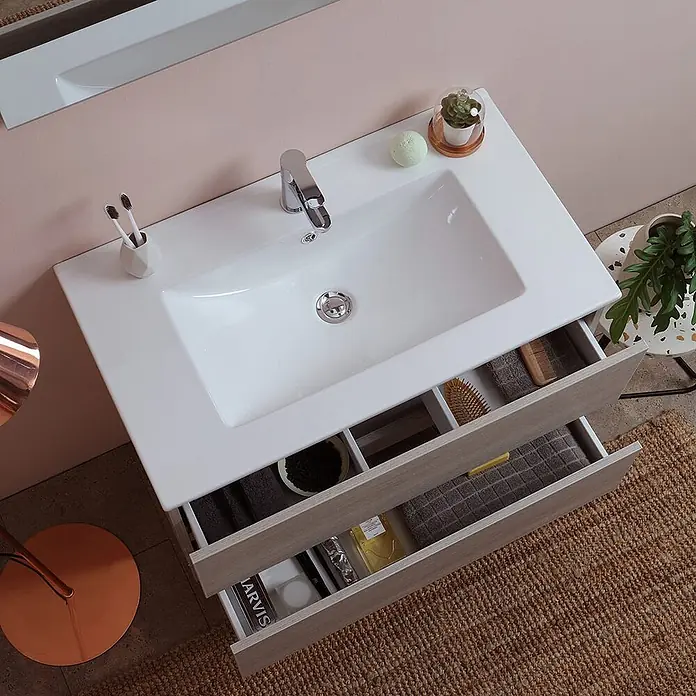 Kiamami Valentina Badmöbel-Set - Helles Holz - Badezimmerschrank mit weißem Keramikwaschbecken und hellen Holzschubladen, teils geöffnet mit Toilettenartikeln.