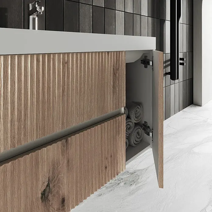 Kiamami Valentina Badmöbel-Set - Braun - Moderne Badezimmer-Waschtischunterschrank mit geriffelten hellen Holzfronten und weißer Arbeitsplatte, offene Tür zeigt gerollte graue Handtücher.