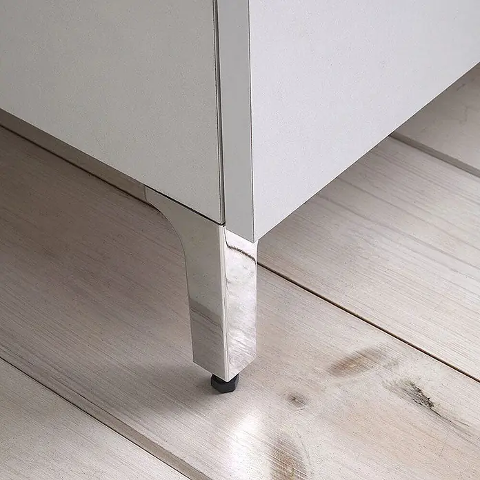 Kiamami Valentina Badmöbel-Set - Weiß - Nahaufnahme eines glänzenden Chrom-Möbelfußes mit schwarzem Stellfuß, der einen weißen Schrank auf hellem Holzboden stützt.