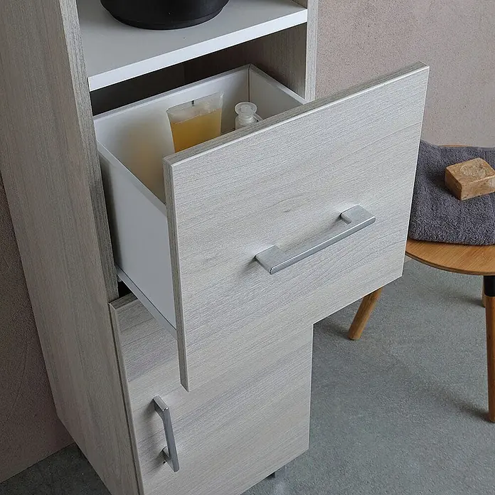 Kiamami Valentina Badmöbel-Set - Grau - Hoher Aufbewahrungsschrank in heller Holzoptik mit geöffneter Schublade, die Toilettenartikel zeigt. Weiße Ablagen und silberne Griffe.