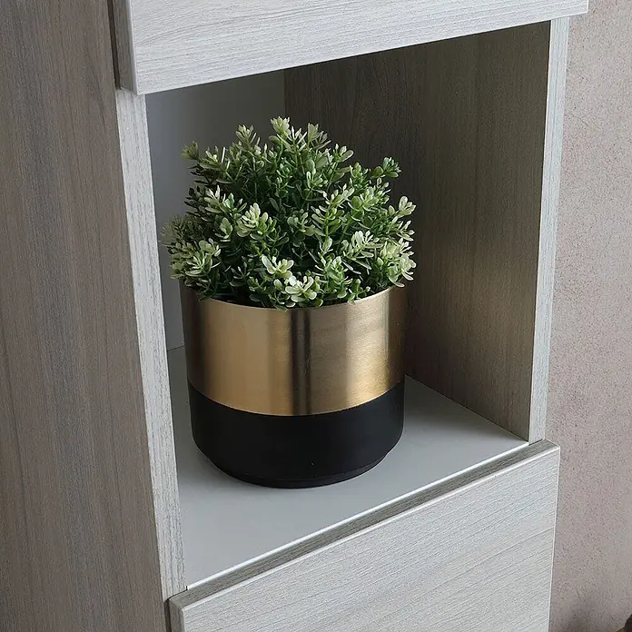 Kiamami Valentina Badmöbel-Set - Grau - Kleine Kunstpflanze in einem gold-schwarzen Pflanzgefäß, auf einem hellen Regal in Holzoptik.