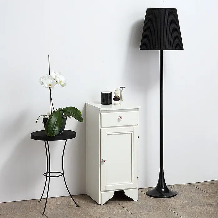 Kiamami Valentina Badmöbel-Set - Weiß - Weißer Nachttisch aus Holz mit einer Schublade und einem Schrankfach, gezeigt in einem Raum mit einer Lampe und Orchidee.