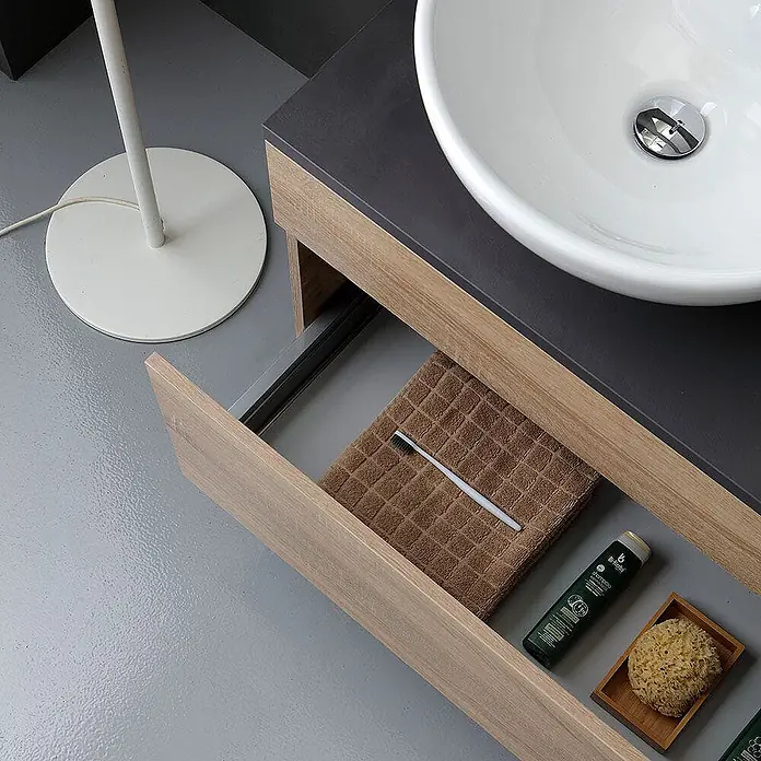 Kiamami Valentina Badmöbel-Set - Helles Holz - Offene Waschtischschublade in modernem Bad, mit Handtuch, Zahnbürste, Shampoo und Schwamm neben weißem Waschbecken.
