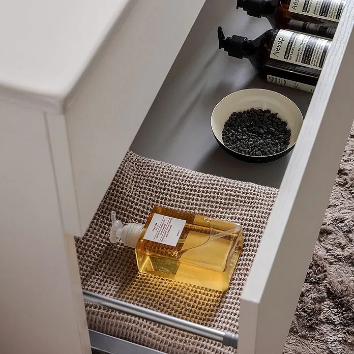 Kiamami Valentina Badmöbel-Set - Weiß - Eine klare, rechteckige Handseifenflasche mit Pumpe auf einer beigen Matte in einer Schublade, mit dunklen Aesop-Flaschen im Hintergrund.