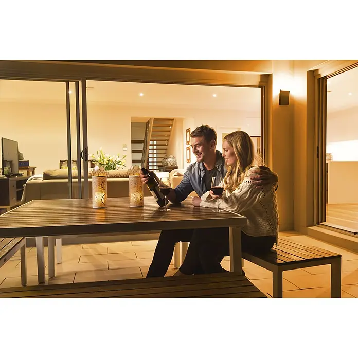Ledvance Smart+ LED-Deckenleuchte rund Curve Dowm RGBW - RGBW - Holztisch und Bänke für den Außenbereich mit hellem Metallrahmen, genutzt von einem Paar, das auf einer Terrasse Getränke genießt.