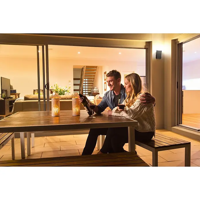 Ledvance Smart+ LED-Deckenleuchte rund Curve Dowm RGBW - RGBW - Zwei dekorative weiße Outdoor-Laternen mit filigranen Mustern, warm leuchtend auf einem hölzernen Terrassentisch, an dem ein Paar nachts entspannt.