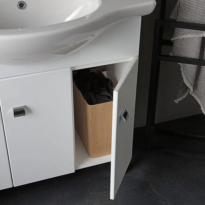 Kiamami Valentina Badmöbel-Set - Weiß - Weißer Badezimmerschrank mit integriertem Waschbecken, offene Tür zeigt Holzkorb mit dunklen Handtüchern.