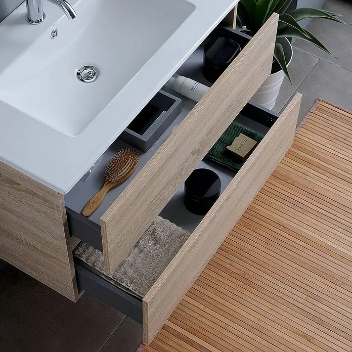 Kiamami Valentina Badmöbel-Set - Braun - Waschtisch aus hellem Holz mit weißem Waschbecken. Offene Schubladen zeigen Haarbürste, Flaschen und Handtücher für praktische Aufbewahrung.