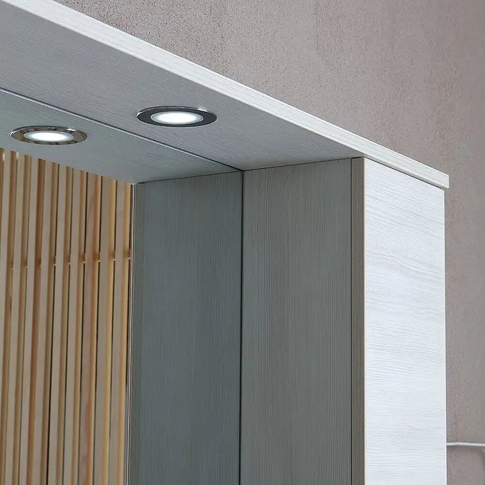 Kiamami Valentina Badmöbel-Set - Weiß - Hell holzgemaserter Schrank mit zwei integrierten LED-Einbauleuchten, der eine Lamellenholzwand im Spiegel reflektiert.