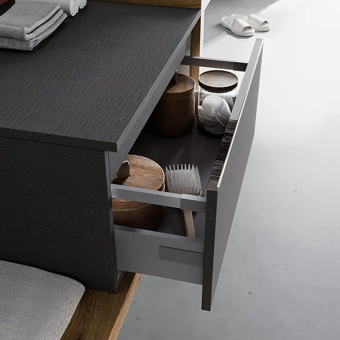 Kiamami Valentina Badmöbel-Set - Grau - Dunkelgrauer strukturierter Badezimmerschrank mit geöffneter Schublade, die Holzdosen, eine Bürste und Wattepads zeigt. Graue Handtücher liegen darauf.