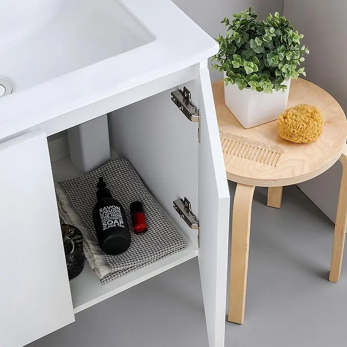 Kiamami Valentina Badmöbel-Set - Weiß - Weißer Bad-Waschtischunterschrank mit offener Tür, der Stauraum zeigt, neben einem hellen Holztisch mit Pflanze, Kamm und Schwamm.