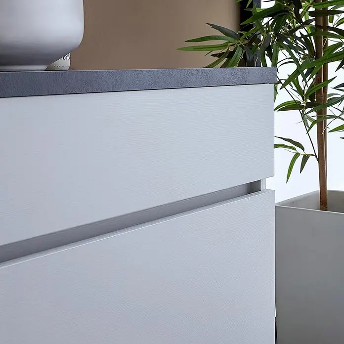 Kiamami Valentina Badmöbel-Set - Weiß - Hellgrauer Schrank in Holzoptik mit zwei Schubladen und dunkler Platte, in modernem Interieur mit Pflanze.