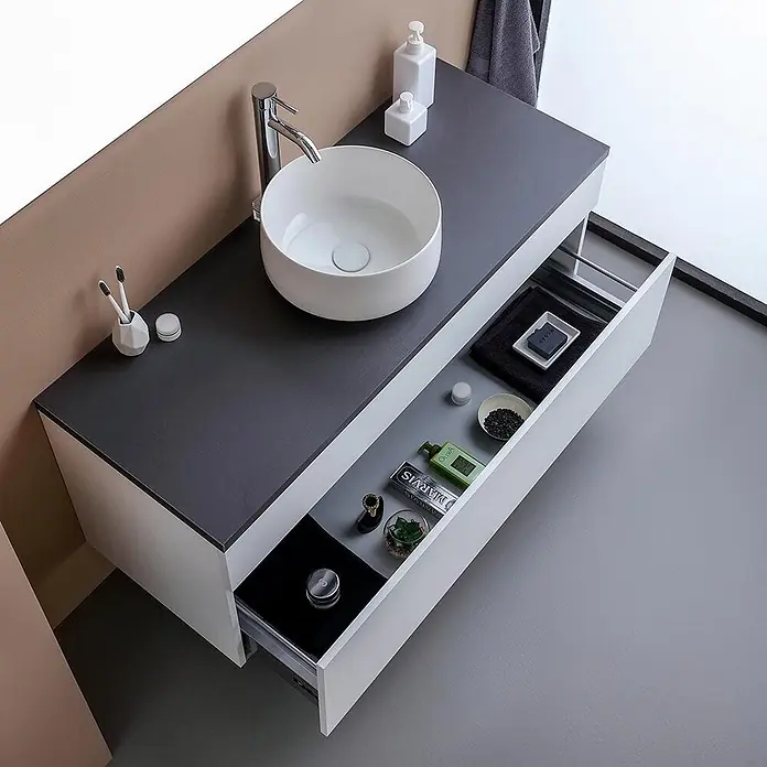 Kiamami Valentina Badmöbel-Set - Weiß - Moderner Waschtisch mit weißem Becken, dunkelgrauer Platte und geöffneter Schublade mit Toilettenartikeln.