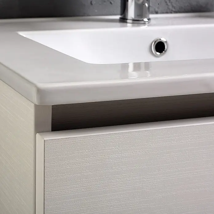 Kiamami Valentina Badmöbel-Set - Weiß - Nahaufnahme eines weißen Keramik-Waschbeckens auf einem hellen Holzmaserung-Badezimmerschrank. Ein Chromwasserhahn und Abfluss sind sichtbar, die Schublade ist leicht geöffnet.
