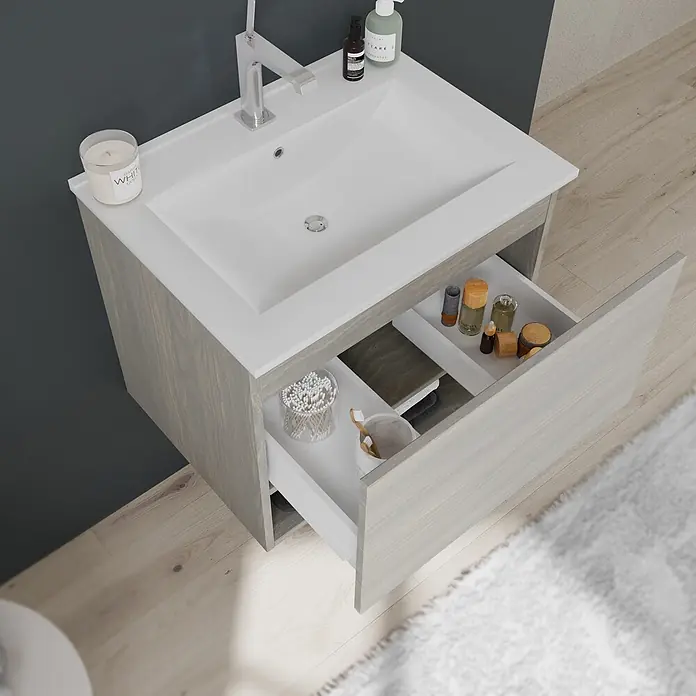 Kiamami Valentina Badmöbel-Set - Helles Holz - Helles graues Holz-Badezimmerwaschtisch mit weißem Waschbecken und Chromarmatur, obere Schublade offen mit Toilettenartikeln.
