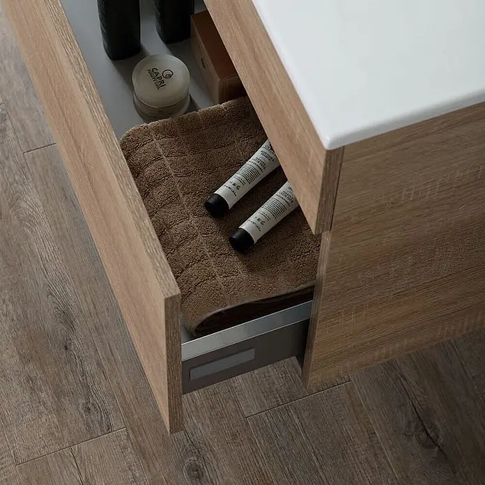 Kiamami Valentina Badmöbel-Set - Helles Holz - Teilweise geöffnete helle Holz-Waschtischschublade mit braunem Handtuch und Toilettenartikeln.