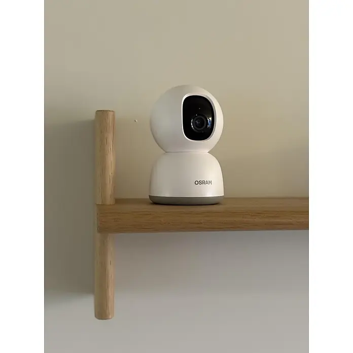 Osram Smart+ Überwachungskamera 360 - Weiß - Weiße OSRAM Überwachungskamera mit schwarzem Objektiv und grauem Sockel auf einem Holzregal.