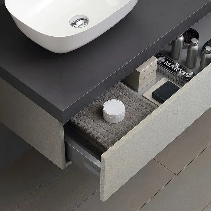 Kiamami Valentina Badmöbel-Set - Grau - Moderne Badmöbel mit dunkelgrauer Arbeitsplatte und hellgrauem strukturiertem Auszug, teilweise geöffnet, zeigt Toilettenartikel und ein Handtuch.