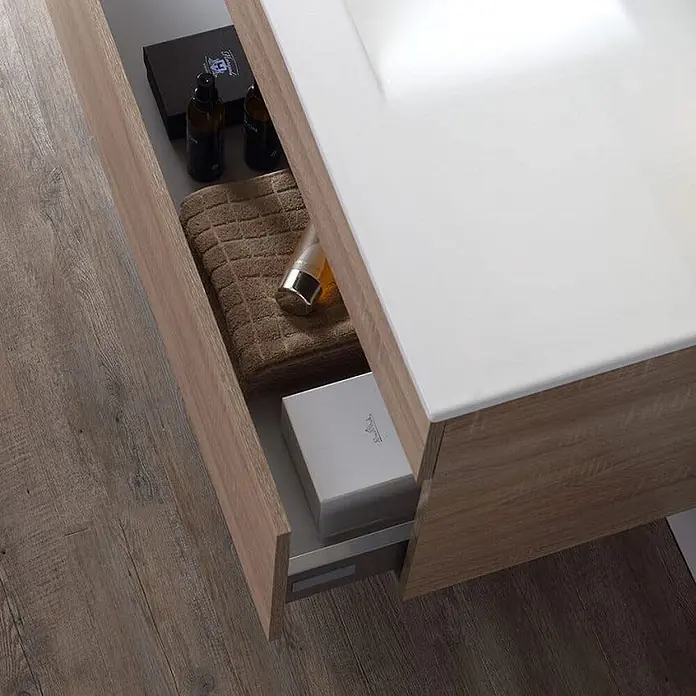 Kiamami Valentina Badmöbel-Set - Helles Holz - Offene Badezimmer-Waschtischschublade in Holzoptik mit weißer Platte, die Toilettenartikel und ein Handtuch enthält.