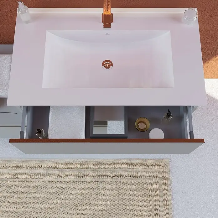Kiamami Valentina Badmöbel-Set - Grau - Weißer Badschrank mit integriertem Waschbecken, Kupferarmatur. Offene Schublade zeigt Handtücher und Accessoires, cremefarbene Badematte.