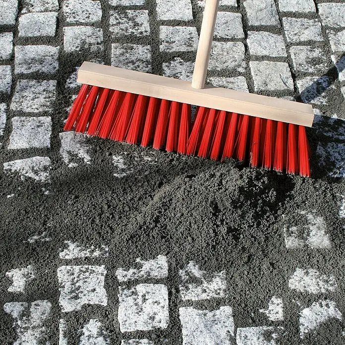 Dansand Steinmehl No Grow - Anthrazit, 20 kg - Nahaufnahme eines Besens mit roten Borsten, der auf einem Pflasterweg mit Sand liegt. Der Besen ist ein Werkzeug zum Reinigen von Oberflächen.