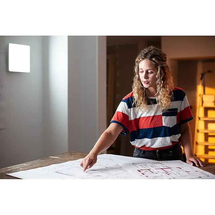 Ledvance Smart+ LED-Wand- & Deckenleuchte Planon Frameless - 20 W, L x B x H: 30 x 30 x 6,2 cm, Neutralweiß - Eine Frau mit lockigem Haar und gestreiftem T-Shirt zeigt auf Baupläne auf einem Holztisch.
