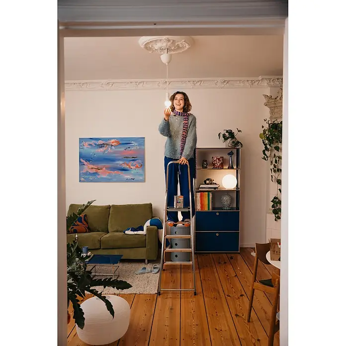 Osram Smart+ Smart-LED-Lampe matter - E27, Dimmbar, RGBW, 1.521 lm, 14 W - Frau auf Leiter installiert eine leuchtende LED-Glühbirne. Bunte Glühbirnenverpackung sichtbar.