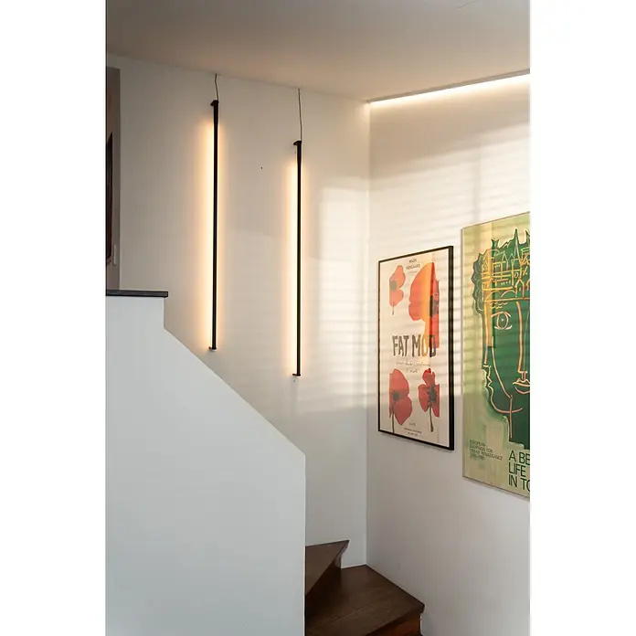 Ledvance Smart+ WiFi LED-Wandleuchte Floor Corner - L x B x H: 142 x 11 x 72 cm, Aluminum, Kaltweiß - Zwei schwarze vertikale Wandleuchten erhellen eine weiße Treppe und Wand, ergänzt durch einen Decken-LED-Streifen. Zwei gerahmte Poster hängen daneben.