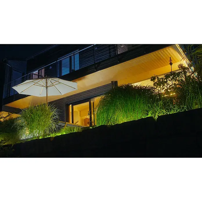 Osram Smart+ LED-Band Flex außen - Länge: 300 cm, Lichtfarbe: RGBW, 12,5 W, 740 lm, 220 V - 240 V - Moderner Hausaußenbereich bei Nacht mit hellem Sonnenschirm, beleuchtetem Holzbalkon und grünen Pflanzen.