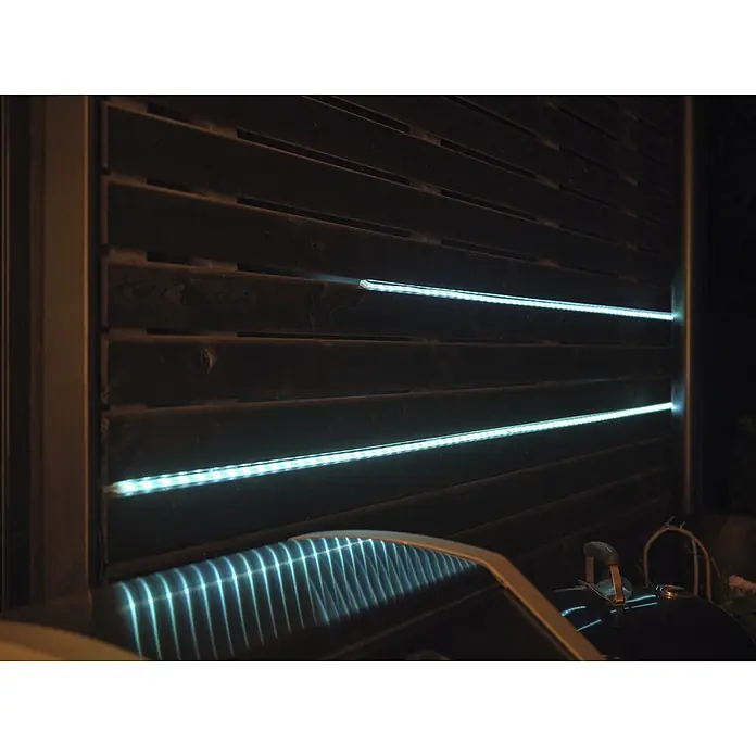 Osram Smart+ LED-Band Flex außen - Länge: 300 cm, Lichtfarbe: RGBW, 12,5 W, 740 lm, 220 V - 240 V - Weiße LED-Streifen beleuchten einen dunklen Holzlattenzaun, mit Reflexionen auf einem Grilldeckel.