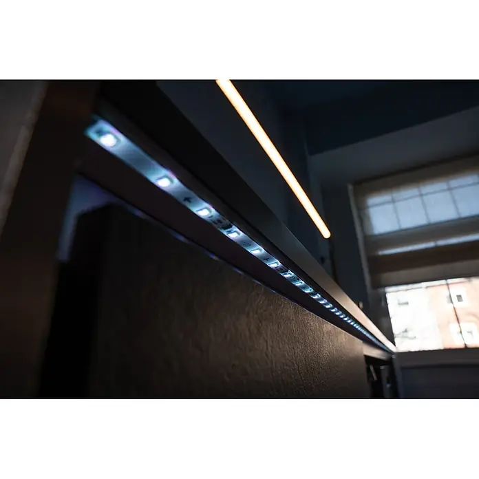 Osram Smart+ LED-Band Flex - Länge: 300 cm, Lichtfarbe: RGB, 15,5 W, 220 V - 240 V - LED-Lichtleiste unter einem dunklen Regal, die eine strukturierte Oberfläche beleuchtet. Eine warme Linienleuchte ist ebenfalls sichtbar.