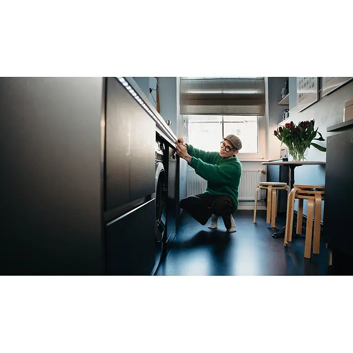 Osram Smart+ LED-Band Flex - Länge: 300 cm, Lichtfarbe: RGB, 15,5 W, 220 V - 240 V - Person stellt eine in dunkle Schränke integrierte Frontlader-Waschmaschine unter Arbeitsplattenbeleuchtung ein.