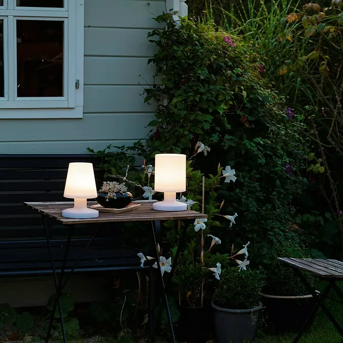 Reality Leuchten Akku-Tischleuchte Lora - 1,5 W, L x B x H: 13 x 13 x 20 cm, Weiß, Warmweiß - Zwei dekorative kabellose Lampen auf einem Tisch im Garten sorgen für stimmungsvolle Beleuchtung.