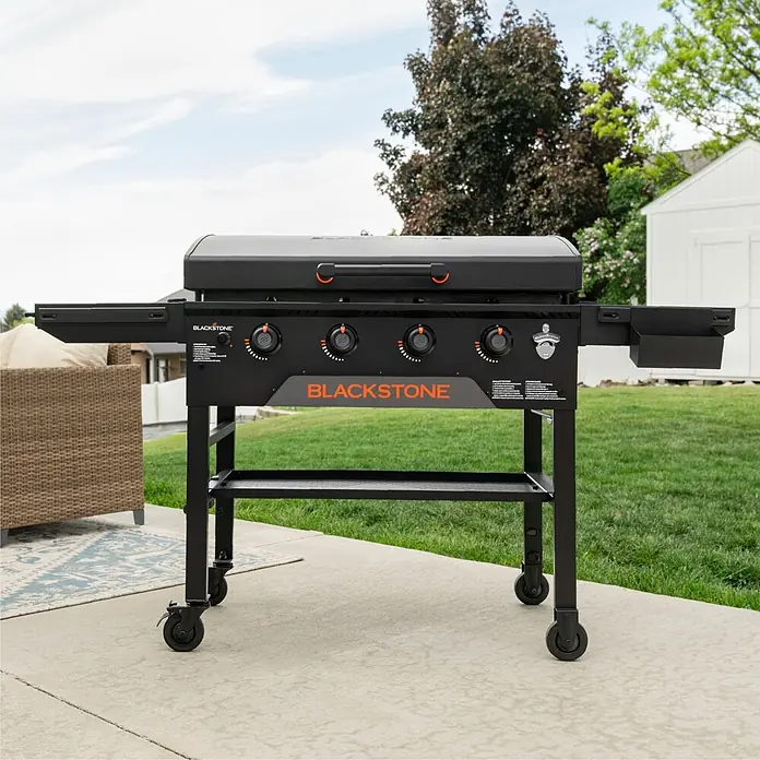 Blackstone Plancha-Grill 36" Omnivore - Grillfläche (B x T): 91 x 55 cm, 11,2 kW, Anzahl Brenner: 4 Stk. - Schwarze Blackstone-Grillplatte mit orangefarbenen Akzenten auf einer Terrasse im Garten, die ihr Design und ihre Größe zeigt.