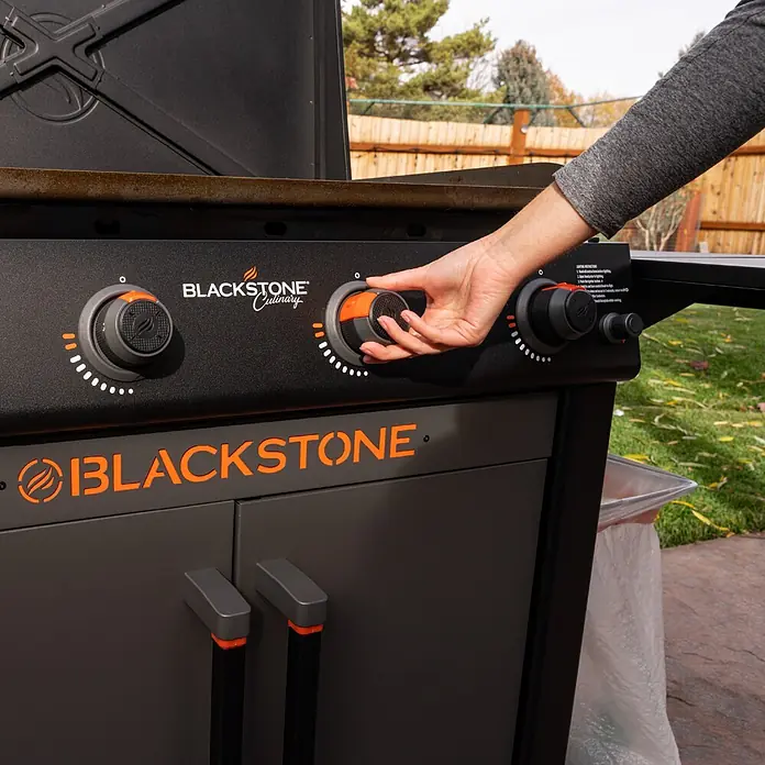 Blackstone Plancha-Grill 36" mit Beistelltisch - Grillfläche (B x T): 91 x 55 cm, 17,6 kW, Anzahl Brenner: 4 Stk., Schwarz - Person verstellt einen Regler an einem schwarzen Blackstone Grill mit orangefarbenen Akzenten im Garten.
