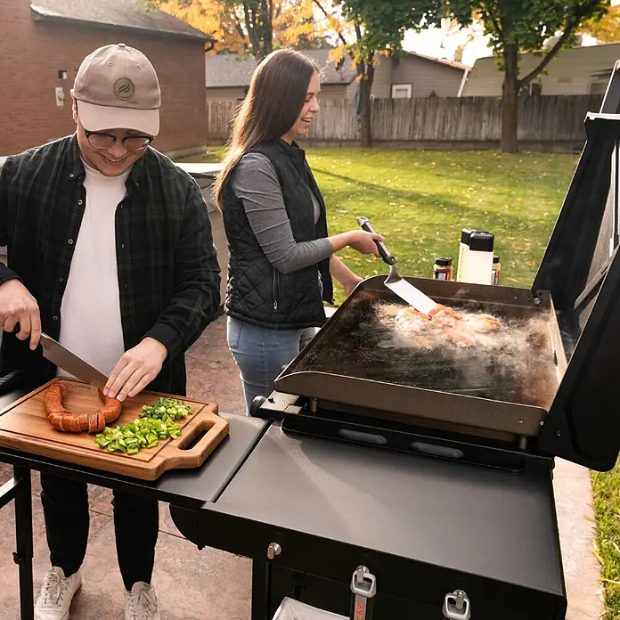Blackstone Plancha-Grill 36" mit Beistelltisch - Grillfläche (B x T): 91 x 55 cm, 17,6 kW, Anzahl Brenner: 4 Stk., Schwarz - Zwei Personen kochen auf einer schwarzen Outdoor-Grillplatte. Einer schneidet Zutaten, der andere brät mit einem Spatel.