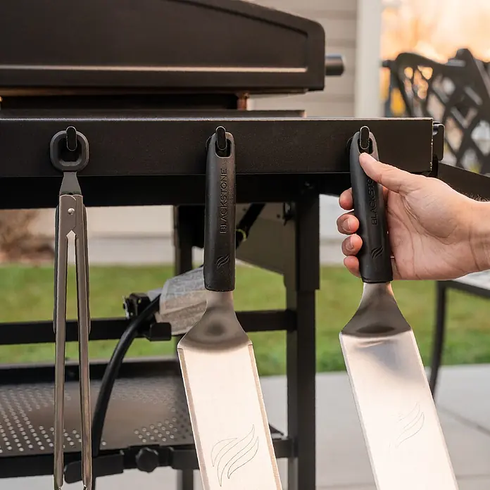 Blackstone Plancha-Grill 28" Omnivore - Grillfläche (B x T): 73 x 45,7 cm, 10 kW, Anzahl Brenner: 2 Stk. - Grillspatel und Zange aus Edelstahl mit schwarzen Griffen, hängend an einem Grill; eine Hand greift nach einem Spatel.