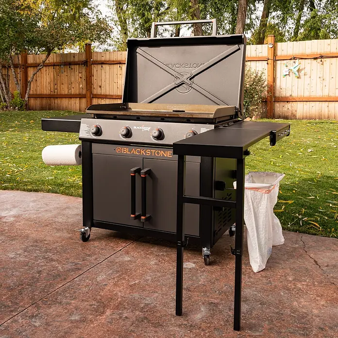Blackstone Plancha-Grill 36" mit Beistelltisch - Grillfläche (B x T): 91 x 55 cm, 17,6 kW, Anzahl Brenner: 4 Stk., Schwarz - Schwarze Blackstone-Außengrillplatte mit offenem Deckel, Kochfläche, Papierhandtuchhalter und Beistelltisch mit Müllsack auf einer Terrasse.
