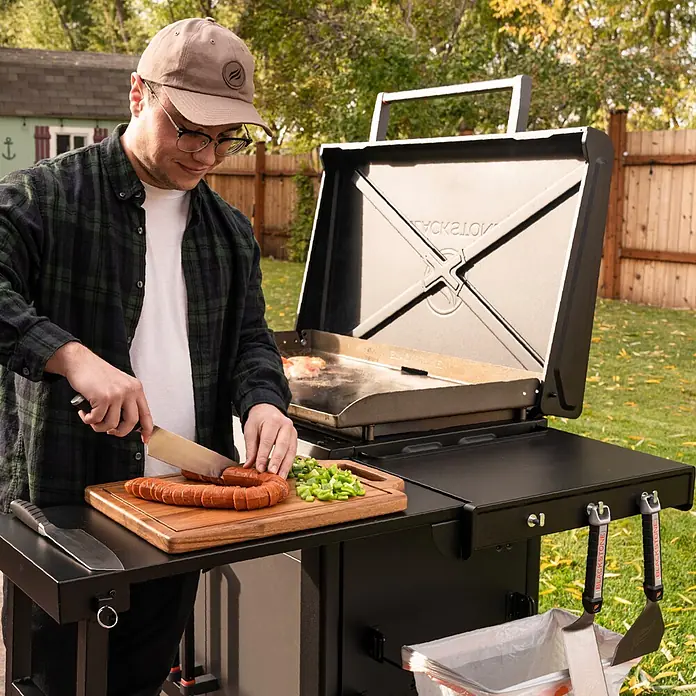 Blackstone Plancha-Grill 36" mit Beistelltisch - Grillfläche (B x T): 91 x 55 cm, 17,6 kW, Anzahl Brenner: 4 Stk., Schwarz - Mann schneidet Wurst auf Holzbrett neben schwarzem Grill mit kochendem Essen und hängenden Spateln im Freien.