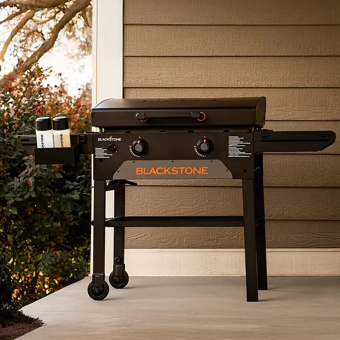 Blackstone Plancha-Grill 28" Omnivore - Grillfläche (B x T): 73 x 45,7 cm, 10 kW, Anzahl Brenner: 2 Stk. - Schwarze Blackstone-Grillplatte mit orangefarbenen Akzenten, Rädern und Gewürzflaschen auf einer Veranda.