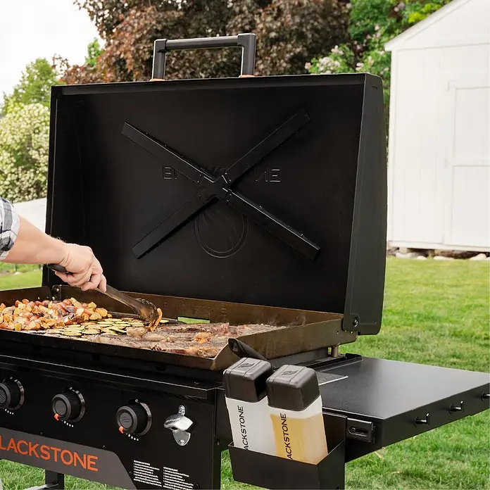 Blackstone Plancha-Grill 36" Omnivore - Grillfläche (B x T): 91 x 55 cm, 11,2 kW, Anzahl Brenner: 4 Stk. - Schwarzer Blackstone-Grill brät Kartoffeln, Zucchini und Fleisch draußen. Eine Person benutzt eine Zange, mit Soßenflaschen daneben.