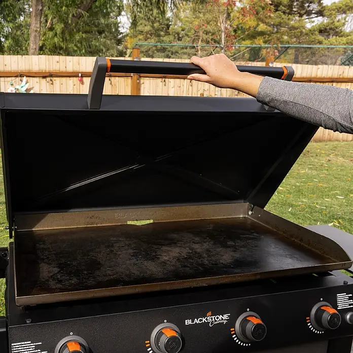 Blackstone Plancha-Grill 36" mit Beistelltisch - Grillfläche (B x T): 91 x 55 cm, 17,6 kW, Anzahl Brenner: 4 Stk., Schwarz - Eine Hand öffnet den schwarzen Deckel eines Blackstone-Grills und zeigt die eingebrannte Kochfläche und das Bedienfeld im Freien.