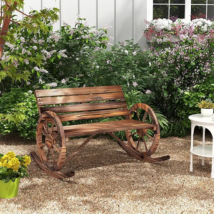 Furnicato Gartenbank karbonisiertes Tannenholz - B x T x H: 88 x 105 x 74 cm, Tannenholz, Verkohlt - Dunkle Holz-Gartenbank mit einzigartigen Wagenrad-Armlehnen, umgeben von Pflanzen und Blumen auf einem Kiesweg.