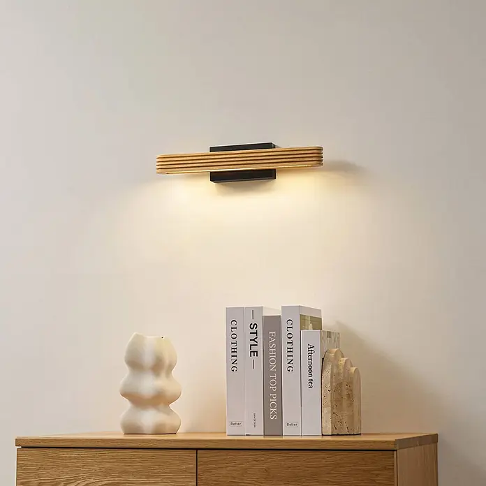 Lucande Wandleuchte Lieske - Holz hell - Moderne LED-Wandleuchte mit schwarzem Sockel und horizontalem Lamellenschirm aus Holz über einem Holzschrank.